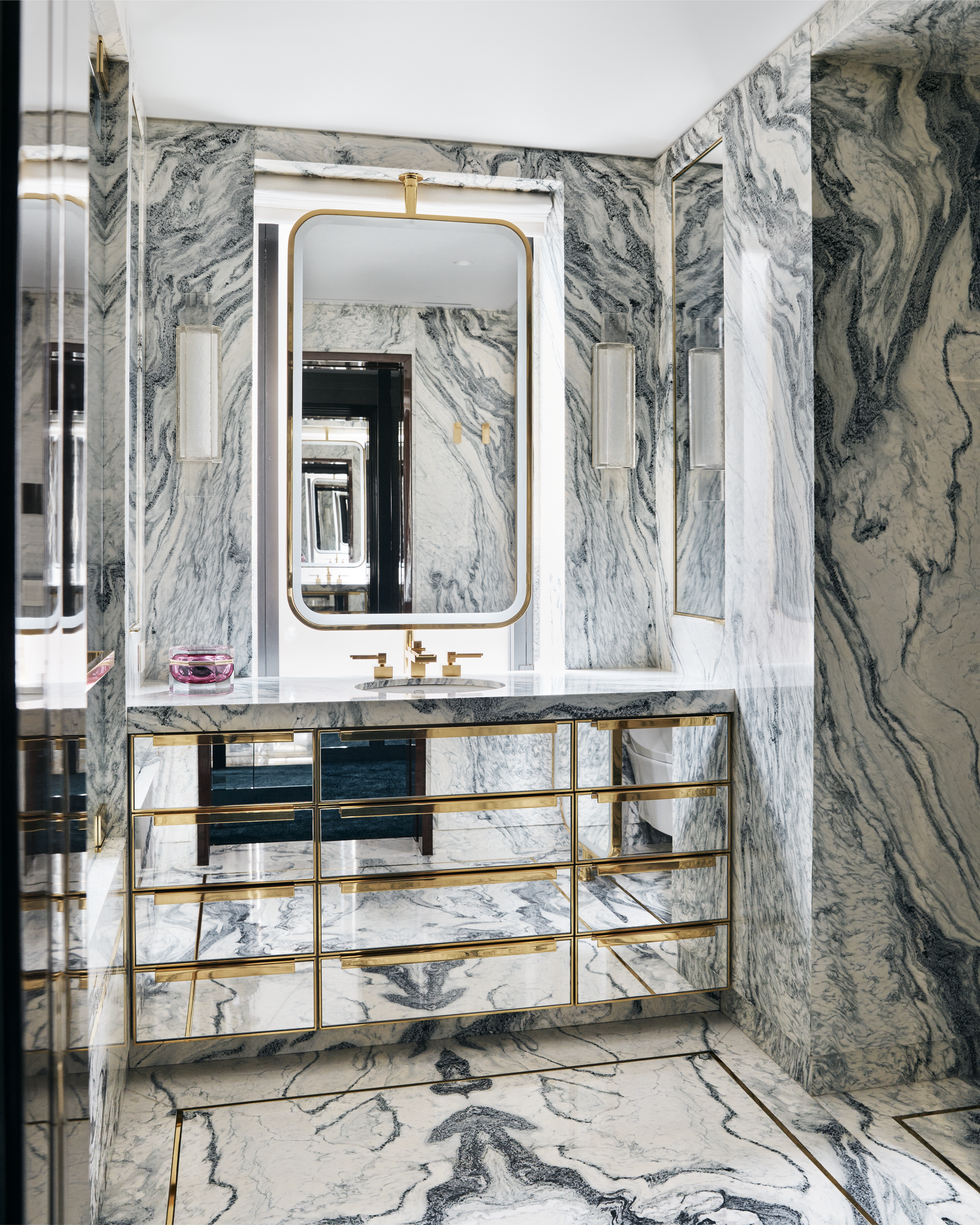 New York City interior design by Pembrooke & Ives. Primary bathroom with marble, mirrored vanity,...